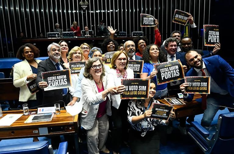Senadores comemoram a aprovação da Lei de Cotas Grupo de senadores celebrando a aprovação da Lei de Cotas no Senado, com cartazes dizendo 'Eu voto sim pela Lei de Cotas'.