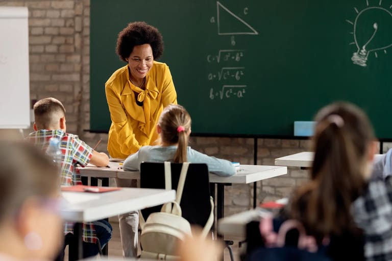 Professora sorrindo e interagindo com alunos em sala de aula, explicando conteúdo em quadro negro.