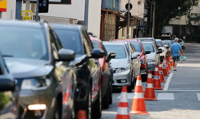Carros parados em fila em frente a um posto de gasolina, com cones de sinalização na calçada.