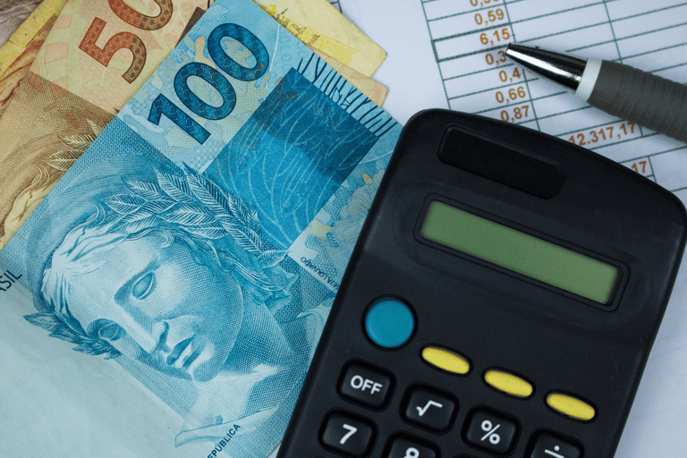 A imagem mostra várias cédulas de real ao lado de uma calculadora preta e uma caneta prata, colocadas sobre uma planilha com números e valores. A cena representa o cálculo financeiro, gerenciamento e planejamento econômico.