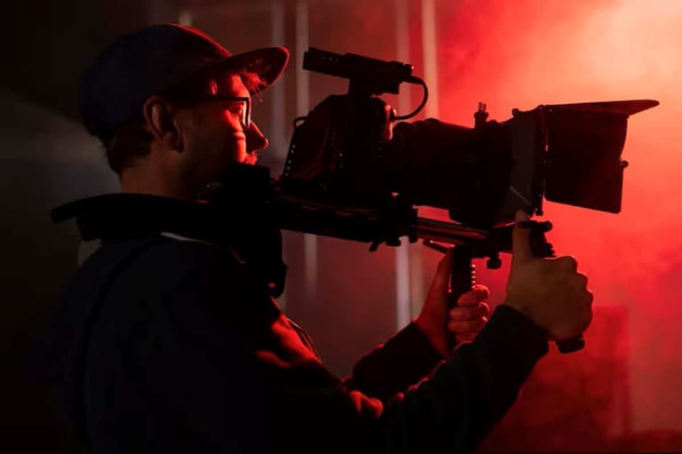 Cinegrafista com boné, filmando com câmera profissional em um set de gravação, iluminado por luz vermelha.