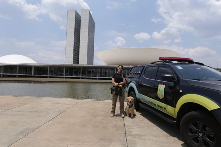 Policial Legislativa Federal ao lado de viatura e cão farejador em frente ao Congresso Nacional em Brasília
