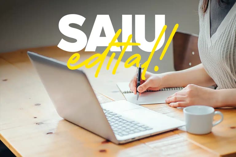 Pessoa escrevendo em caderno ao lado de notebook em mesa de madeira, com texto SAIU Edital em branco e amarelo.