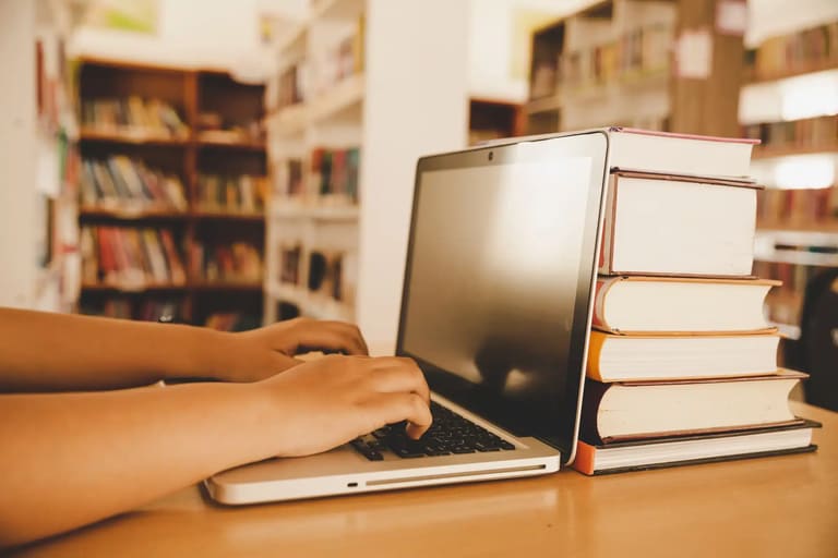 Pessoa utilizando notebook em biblioteca para acompanhar processos seletivos na área da Educação com inscrições abertas.