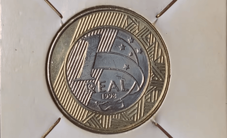 Fotografia em macro de uma moeda de 1 real brasileira do ano de 1998, centralizada em um suporte de papel branco (coin holder). A imagem destaca o número "1", a palavra "REAL" e o ano "1998" gravados no núcleo prateado, rodeado pelo anel dourado com grafismos indígenas marajoaras e estrelas do Cruzeiro do Sul.