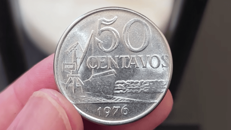 Close-up do reverso de uma moeda de 50 centavos de 1976, segurada por dedos em primeiro plano. O design apresenta o valor "50 CENTAVOS", o ano "1976" na base e a imagem de um navio cargueiro navegando, com detalhes nítidos do metal prateado.