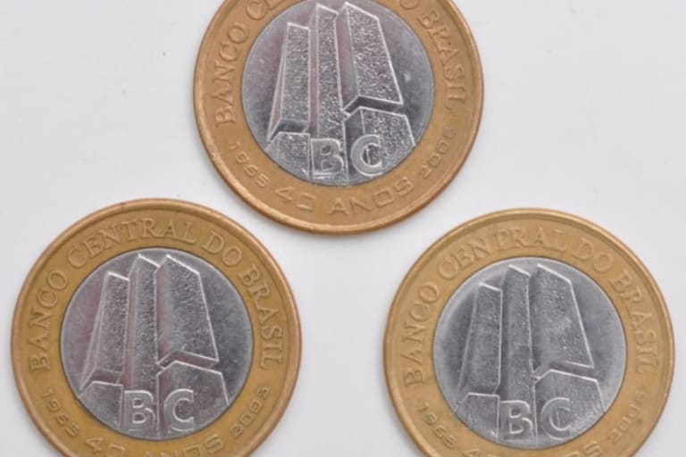 Fotografia de três moedas de 1 Real comemorativas dos 40 anos do Banco Central, organizadas em formato de triângulo sobre um fundo branco. A imagem exibe o lado do anverso das moedas, destacando os detalhes do núcleo prateado e as legendas douradas.