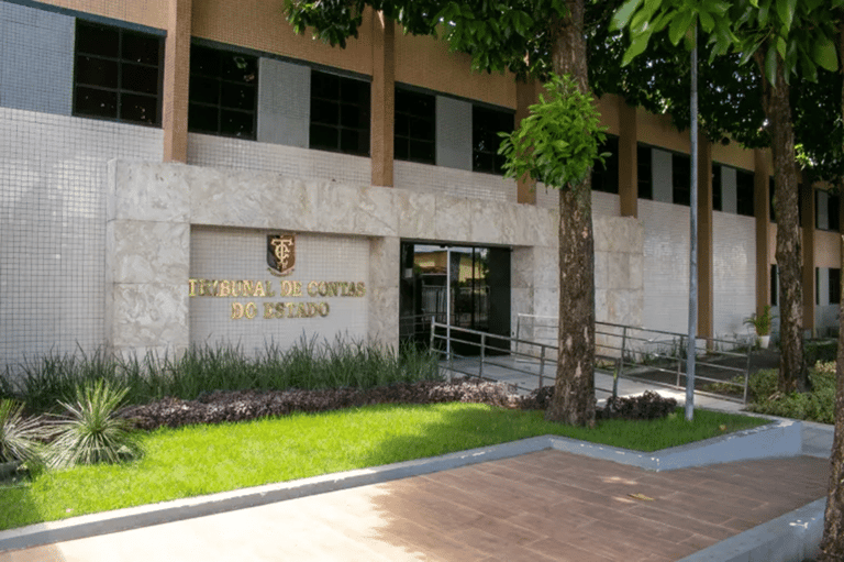 Vista externa do prédio de um Tribunal de Contas do Estado com fachada clara, árvores ao redor e placa em relevo dourado