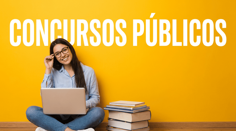 Jovem sorridente de óculos e camisa azul estuda com notebook no colo, ao lado de pilha de livros, fundo amarelo.