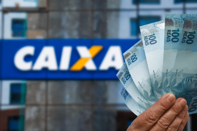Mão segurando notas de R$ 100 em frente a agência da Caixa representando depósito de R$ 7.300 do Auxílio Reconstrução