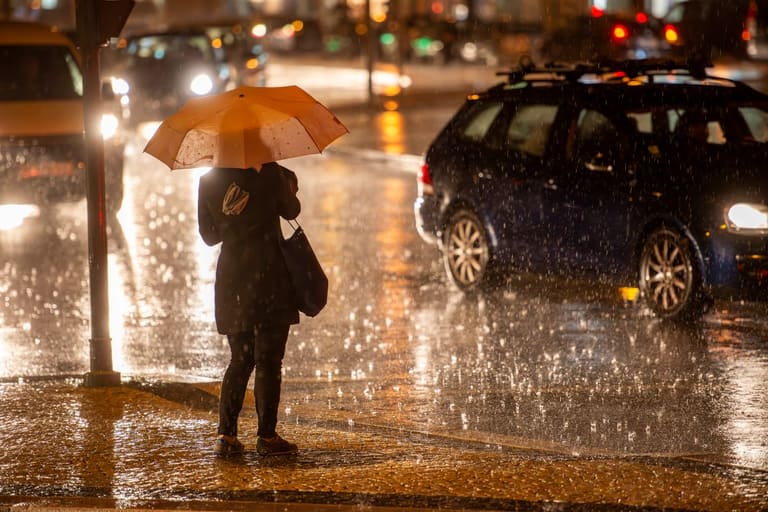 Pessoa com guarda-chuva atravessando rua durante chuva intensa à noite em área urbana.