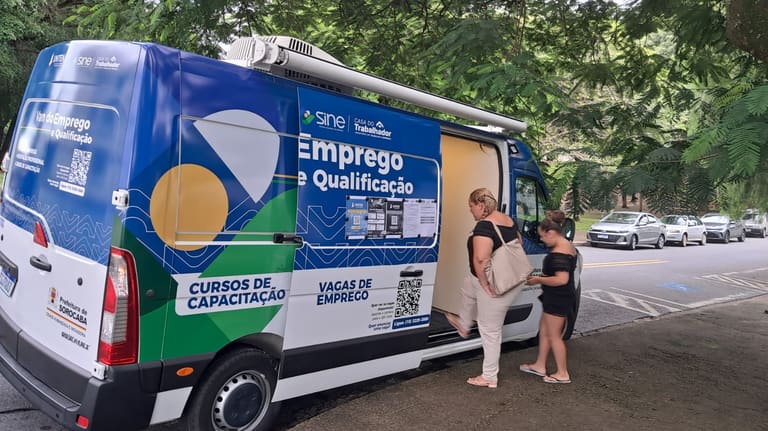 Van itinerante azul do SINE Casa do Trabalhador estacionada em área arborizada com duas mulheres no atendimento