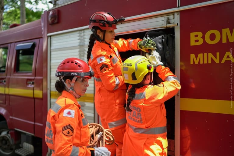 Bombeiras militares de Minas Gerais com uniforme operacional ajustam equipamentos ao lado de viatura
