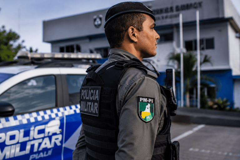 Policial da PM-PI com colete tático preto e boina observa área externa em frente a viatura azul e branca.