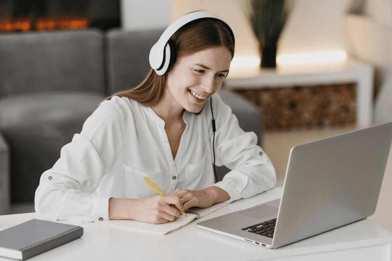 Mulher ruiva sorridente com fone branco faz anotações em caderno enquanto olha para laptop em ambiente doméstico.
