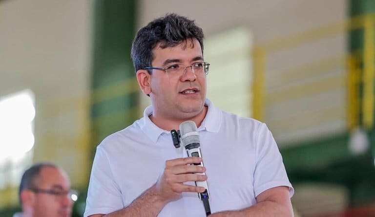 Rafael Fonteles, governador do Piauí desde 2023 , de camisa polo branca e óculos, segura microfone durante discurso em ginásio
