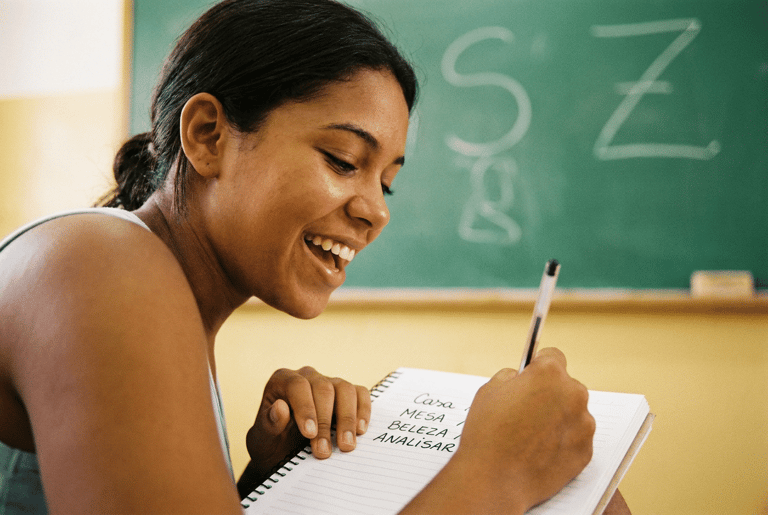 Estudante sorridente escreve lista de palavras em caderno espiral diante de quadro verde com as letras S e Z grafadas a giz