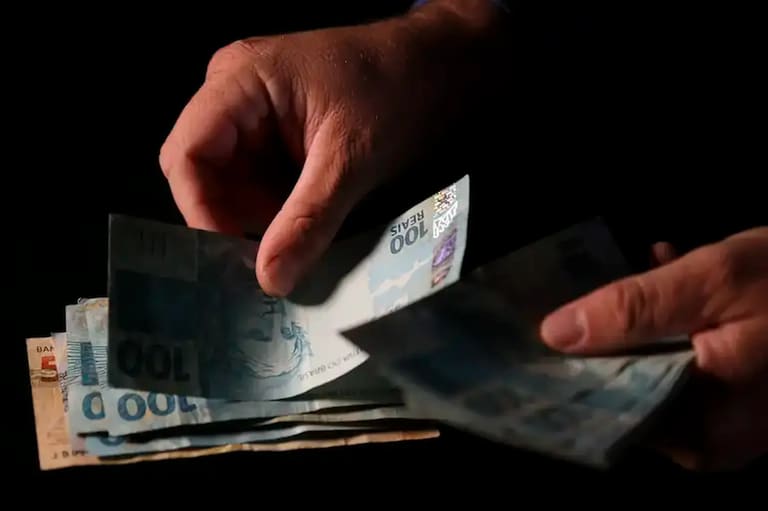 Mãos seguram notas de R$ 100 em imagem sobre pagamento do Auxílio Reconstrução pela Caixa.