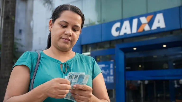 Mulher segura cédulas de reais em frente a uma agência da Caixa, representando os pagamentos do Bolsa Família de junho.