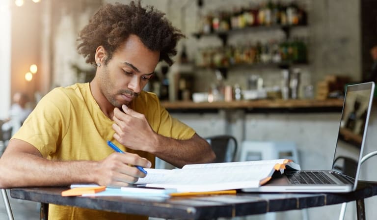 Jovem concentrado estuda apostila e faz anotações ao lado do notebook em preparação para concurso público.