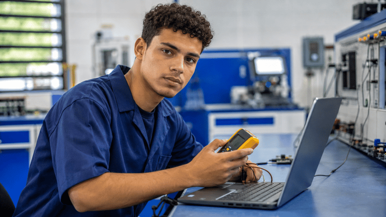 Jovem com cabelo cacheado usa multímetro em laptop em bancada de laboratório tecnológico.