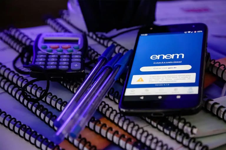 Celular com tela do Enem sobre cadernos e materiais escolares, representando o novo prazo para atendimento especializado e nome social em 2026.