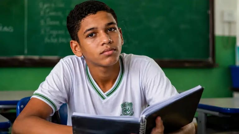 Estudante brasileiro com caderno em sala de aula pública, representando regra de frequência para recebimento do benefício mensal do Pé-de-Meia.