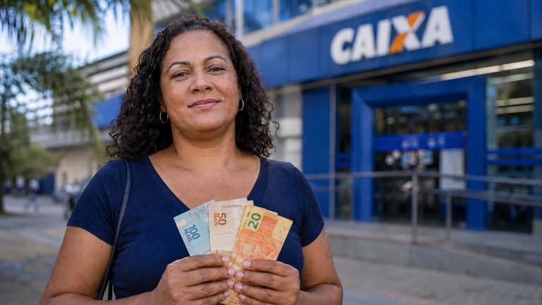 Mulher segura cédulas de real em frente a uma agência da CAIXA, representando a liberação do saque de R$ 6.220.
