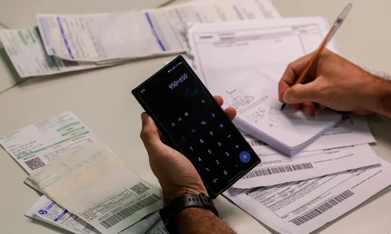 Pessoa calcula valores em celular enquanto organiza boletos e documentos de dívidas sobre a mesa.