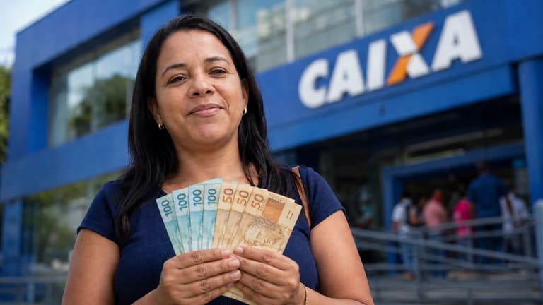Mulher segura notas de real em frente a uma agência da Caixa, representando a confirmação do pagamento de R$ 800.