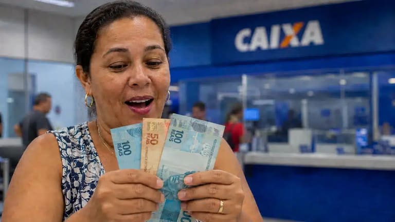 Mulher segura cédulas de real em agência da Caixa, representando o pagamento de R$ 2.900.
