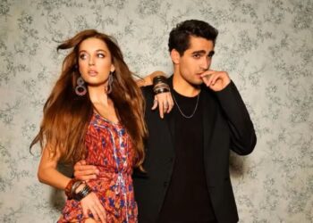 Um casal de atores turcos posando para foto: a mulher à esquerda usa um vestido floral escuro e o homem à direita veste um terno preto com camiseta preta. No centro, uma faixa amarela e preta contém o texto "5 COISAS PROIBIDO NAS NOVELAS TURCAS".