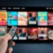 Tela de TV exibindo várias opções de filmes e séries de streaming, com controle remoto em mãos, simbolizando a comparação entre os serviços de Netflix, Max e Prime.