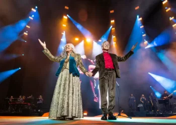 Dois artistas brasileiros se apresentam juntos em um palco iluminado, de mãos dadas, durante um espetáculo musical com banda ao fundo.