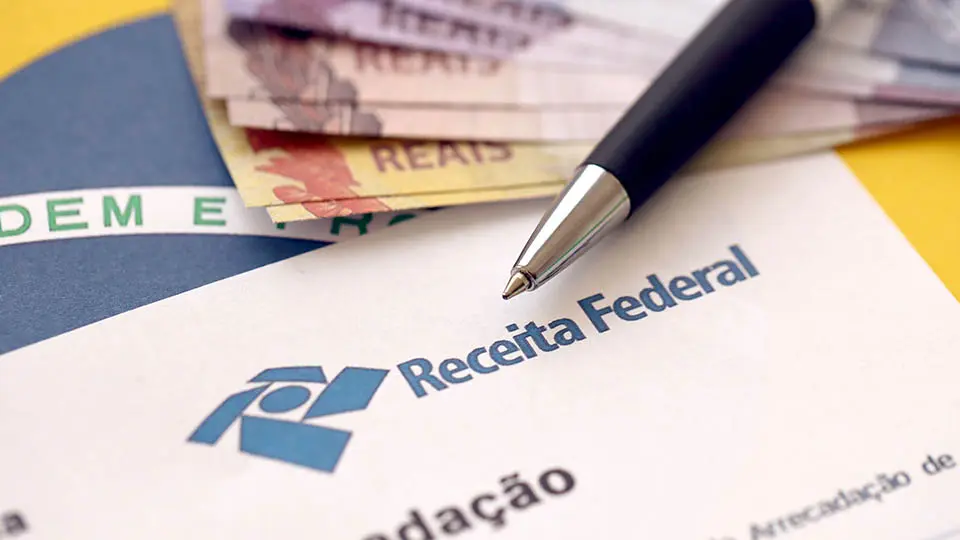Quem ganha até R$ 5 mil receberá alerta da Receita no WhatsApp sobre isenção do IR 2 Caneta sobre documento da Receita Federal com notas de real ao fundo detalhando comunicações oficiais
