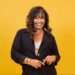 A atriz Solange Couto sorri frontalmente contra um fundo amarelo vibrante. Ela possui cabelos castanhos ondulados com luzes, veste um blazer preto aberto sobre um top de pérolas brancas e usa diversos braceletes dourados no pulso direito.