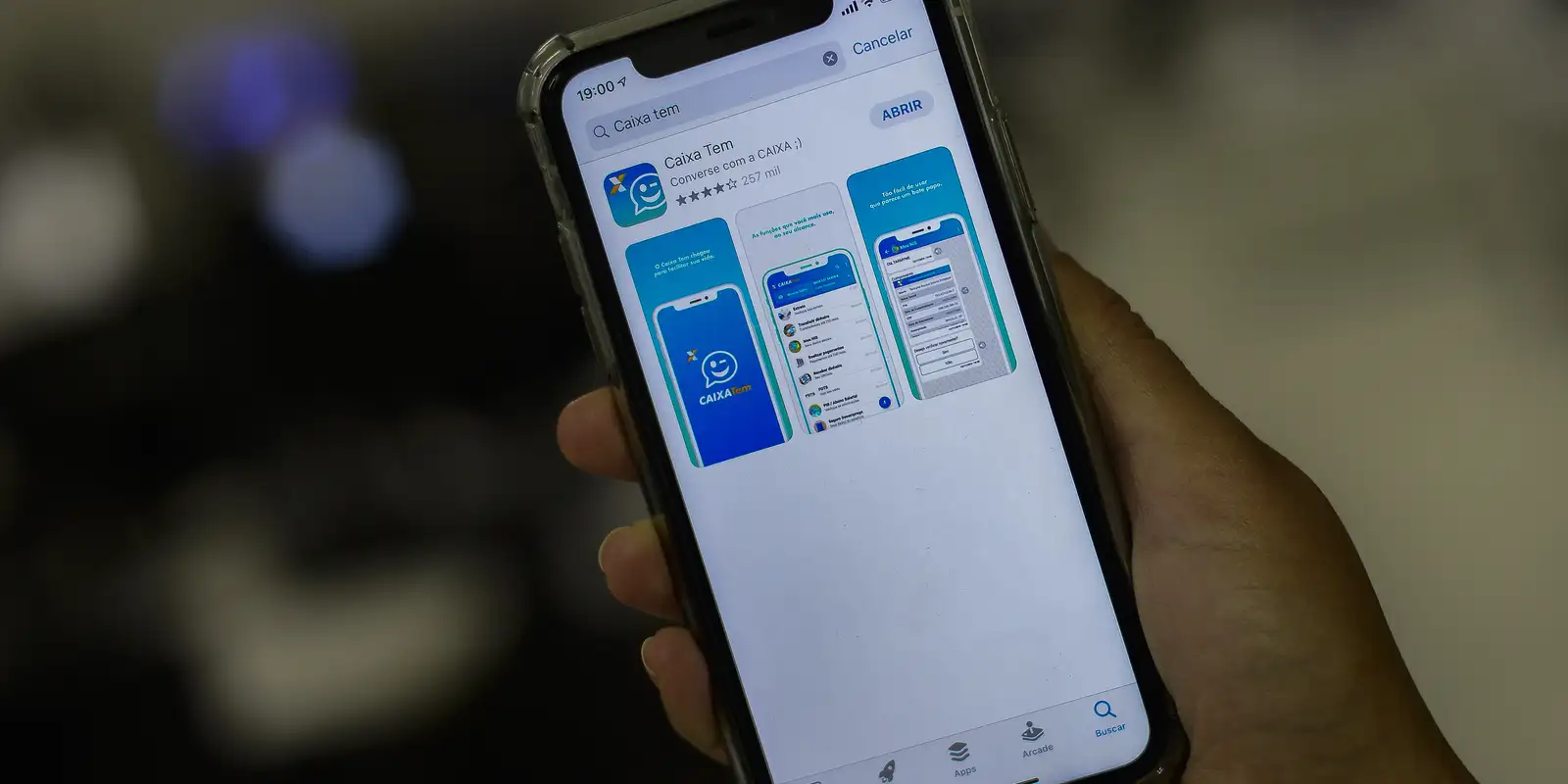 Auxílio Pé-de-Meia 2026: aprenda como fazer o saque do seu benefício 2 Mão segurando celular exibindo tela de download do app Caixa Tem no Brasil