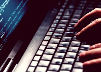 Mãos de pessoa digitando rapidamente em teclado de notebook com tela exibindo linhas de código, exemplificando ataques e golpes, através de aplicativos falsos