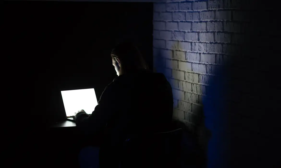 Alerta de segurança: Se você tem este aplicativo instalado, seu banco pode estar em risco 2 Pessoa sentada diante de notebook iluminado, operando no escuro próximo a parede de tijolos, representando ataques a usuários de aplicativos de segurança