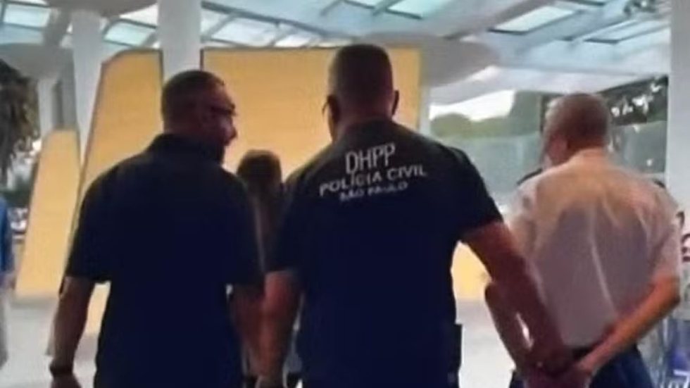 Piloto é preso dentro de avião suspeito de liderar rede de abuso infantil 2 Policiais civis escoltam piloto algemado dentro de aeroporto com ambiente moderno e vidros ao fundo