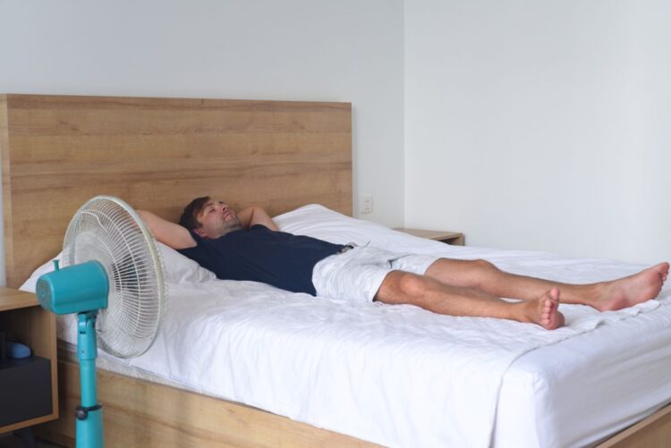 Onda de calor: O ventilador pode causar risco aos pulmões? 1 Homem deitado em uma cama de casal branca, com os braços atrás da cabeça, posicionado à frente de um ventilador de mesa azul ligado, em um quarto com cabeceira de madeira.