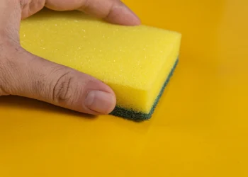 Esponja amarela sendo segurada, com o foco na parte da esponja e fundo amarelo, ilustra o truque das lavanderias sobre o uso de esponjas na máquina de lavar.