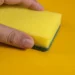 O que acontece se você colocar uma esponja dentro da máquina de lavar? O truque que as lavanderias não contam 13 Esponja amarela sendo segurada, com o foco na parte da esponja e fundo amarelo, ilustra o truque das lavanderias sobre o uso de esponjas na máquina de lavar.