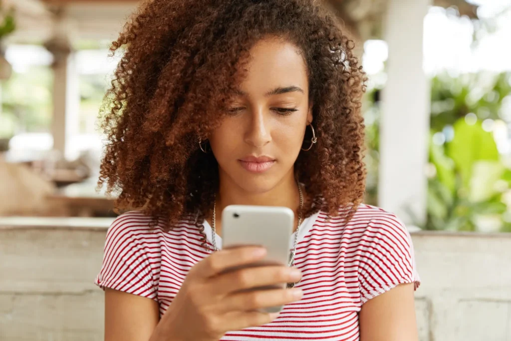 Psicologia do 'Ghosting': Por que algumas pessoas desaparecem sem dar explicações e o que isso revela sobre a maturidade 2 Mulher afro-americana usando o celular em um café, refletindo sobre o impacto emocional do 'ghosting'.