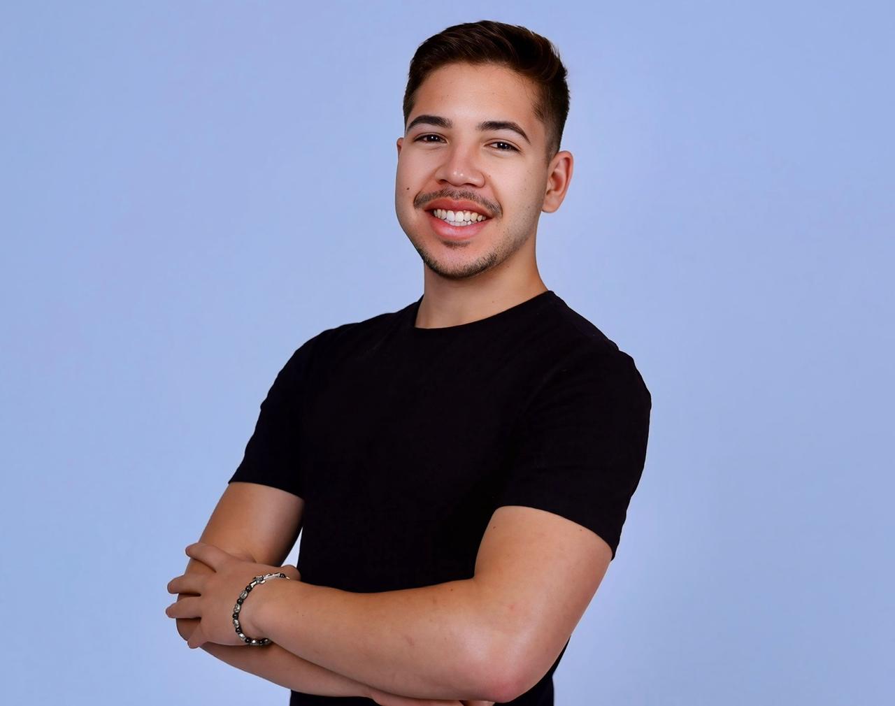 Quer comprar a casa própria? Veja dicas da Caixa e realize seu sonho em 2026 3 Rapaz de camiseta preta sorri com braços cruzados em fundo azul claro