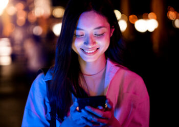 Mulher jovem sorrindo ao enviar mensagem no celular durante a noite com luzes desfocadas ao fundo