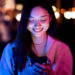 Mulher jovem sorrindo ao enviar mensagem no celular durante a noite com luzes desfocadas ao fundo