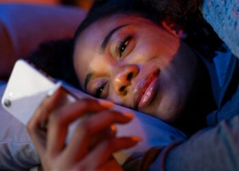 Mulher jovem deitada na cama olhando celular com iluminação noturna em tons de azul e roxo.