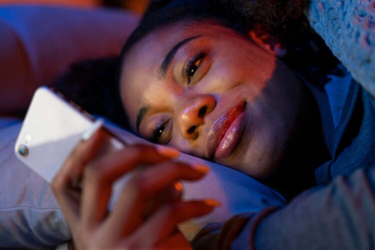 Boa Noite: Mensagens de GRATIDÃO e PAZ para uma noite de sono tranquila 1 Mulher jovem deitada na cama olhando celular com iluminação noturna em tons de azul e roxo.