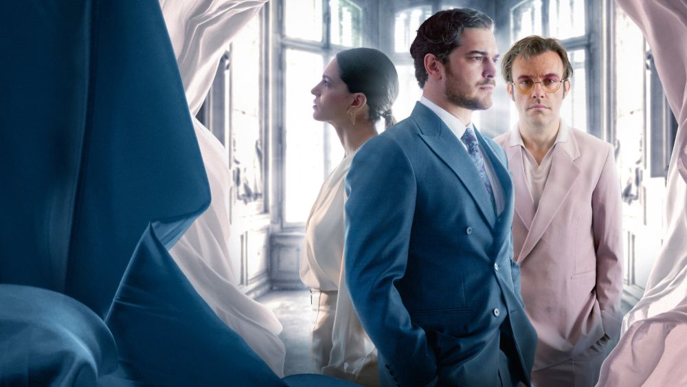 Netflix: 5 séries turcas muito boas que valem a pena assistir 3 Três personagens de O Famoso Alfaiate em um cenário elegante. À frente, um homem de terno azul; ao fundo, uma mulher de perfil e outro homem de terno rosa claro. Tecidos azuis e brancos moldam as laterais da cena.